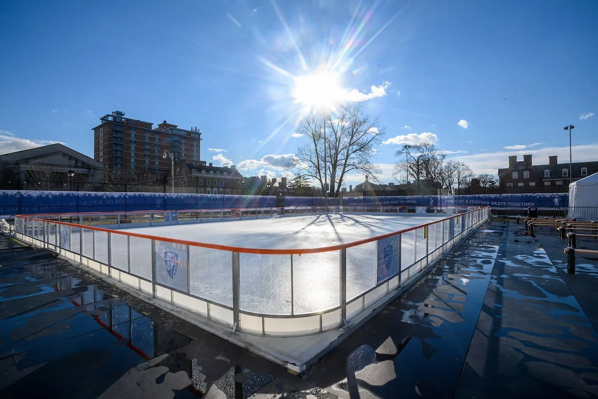 An outdoor ice skating rink reflecting sunlight, surrounded by a metal fence with orange trim, set against a cityscape with buildings and leafless trees, under a bright blue sky.