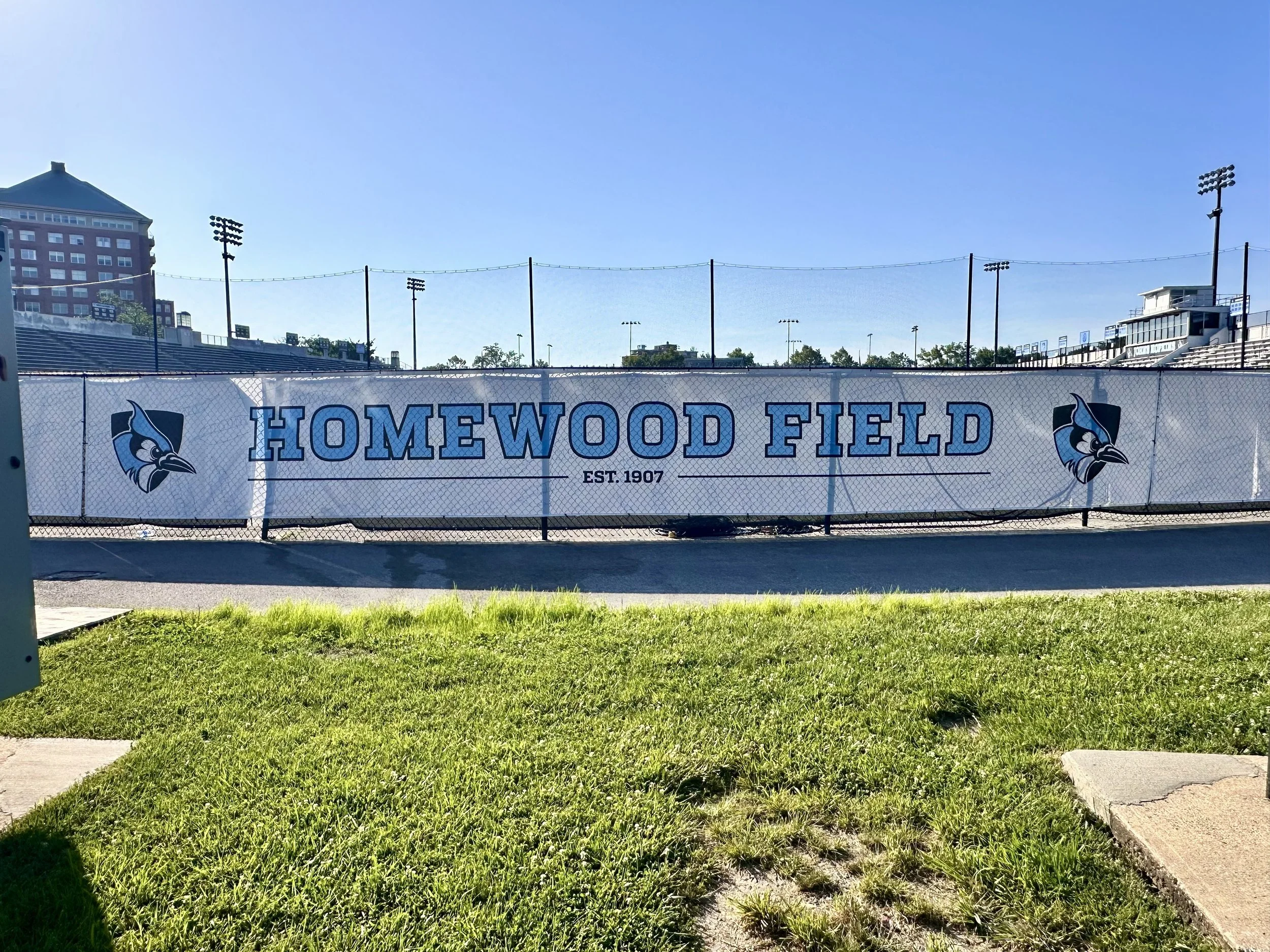 A banner on a chain-link fence reading 'Homewood Field' with an eagle logo on each side, on a sunny day with a clear blue sky at a sports stadium.