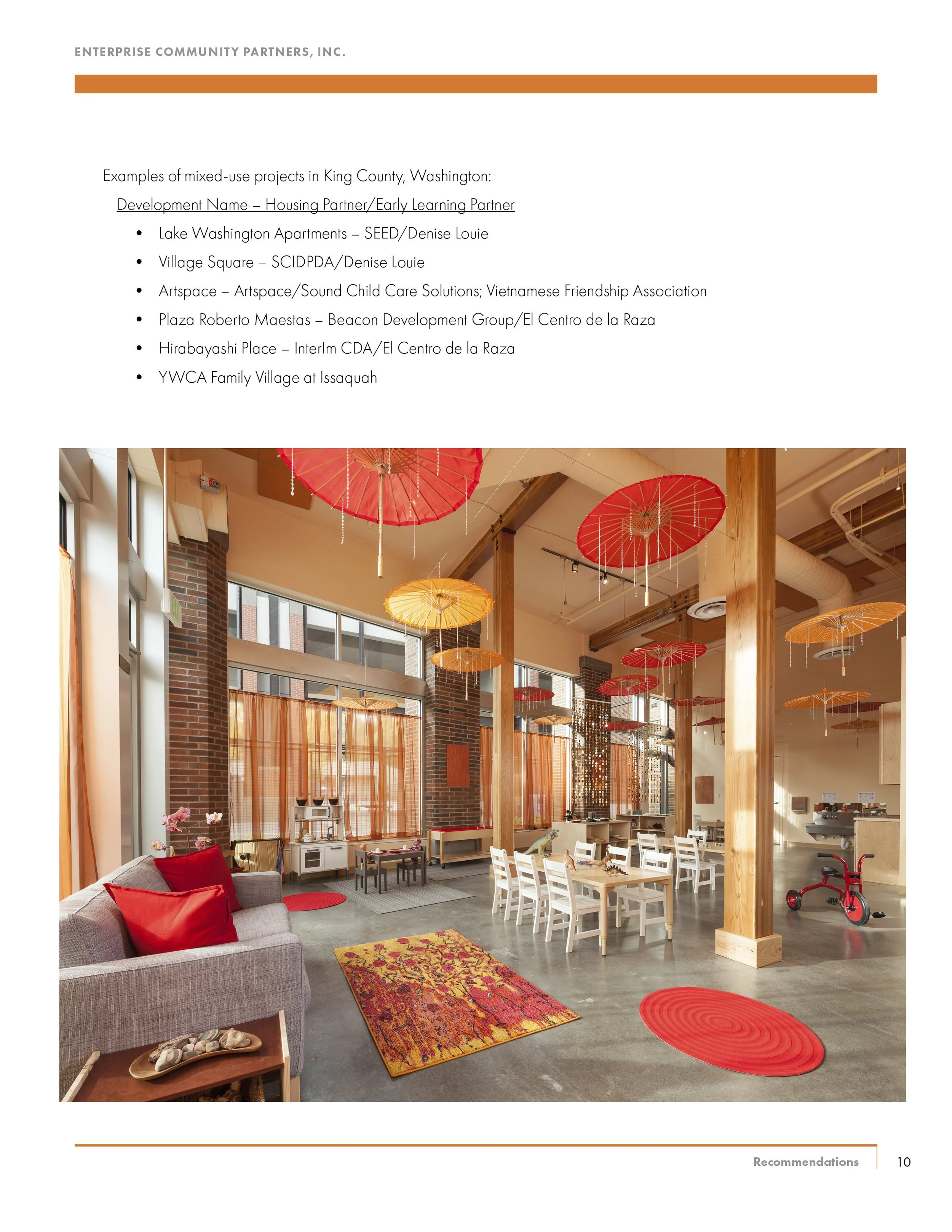 Interior of a community space with hanging red and orange umbrellas, a sofa with red pillows, a patterned rug, white chairs around tables, and large windows with orange curtains.