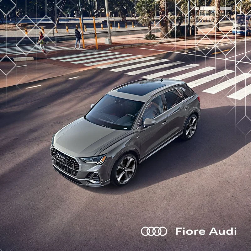 A silver Audi SUV driving on a city street with a crosswalk, seen from an aerial view. The image features a geometric overlay pattern and the text 'Fiore Audi' with the Audi logo in the bottom right corner.