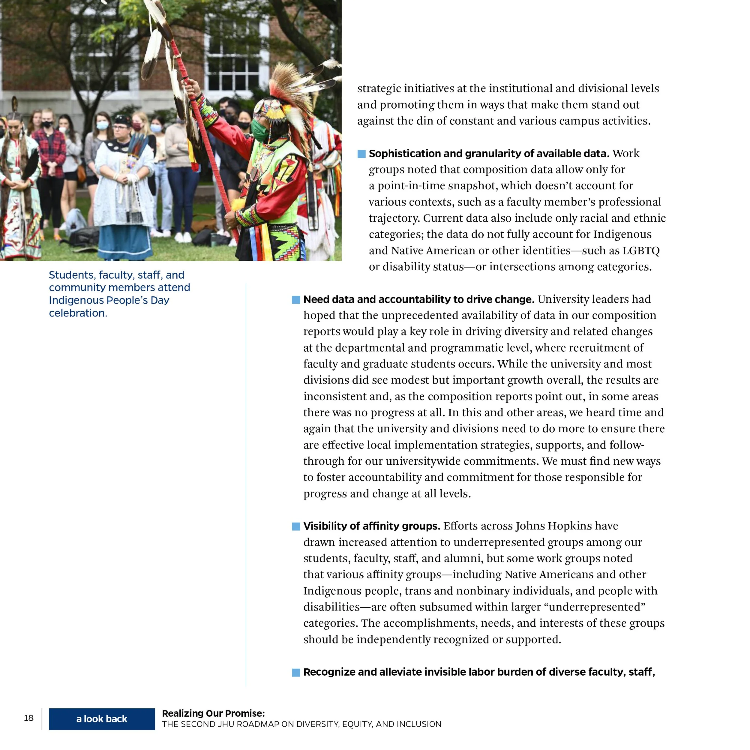 Students, faculty, staff, and community members attending Indigenous People's Day celebration, outdoors on a grassy area with trees, some wearing traditional indigenous attire and face masks.