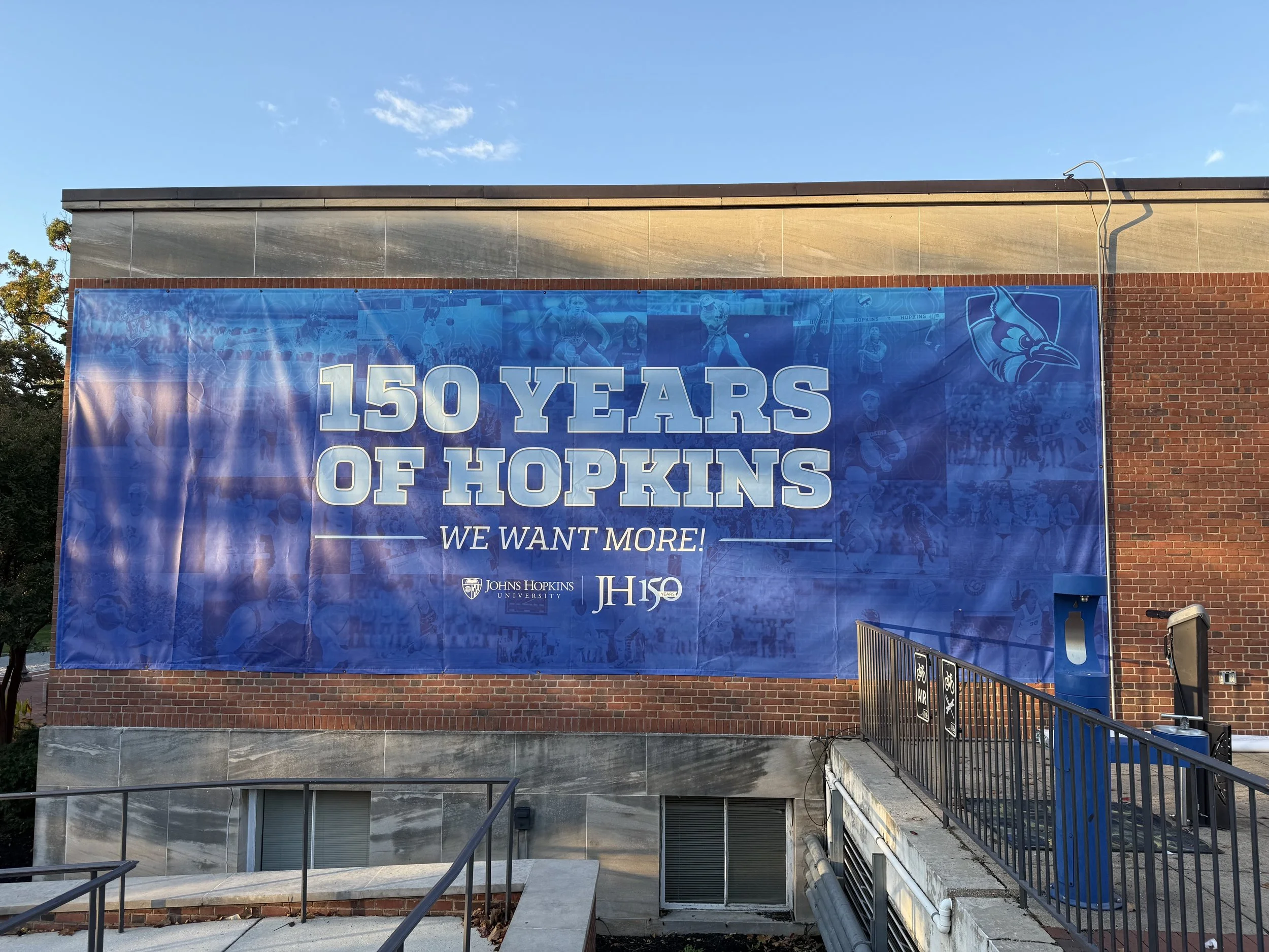 Blue banner on a brick building celebrating 150 years of Johns Hopkins University with the slogan "We Want More!"