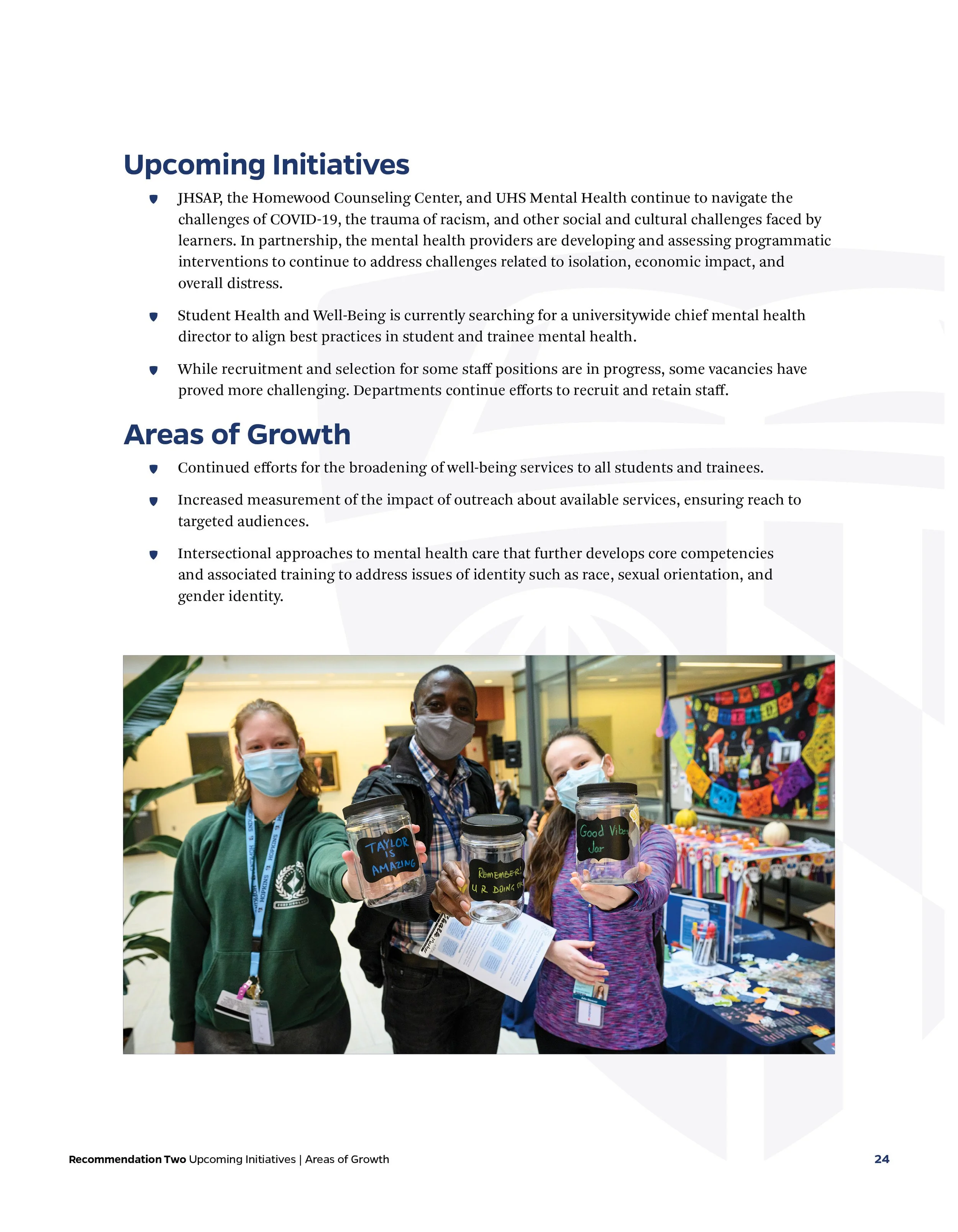 Three people wearing masks holding jars with messages at an indoor event, with a colorful display in the background.