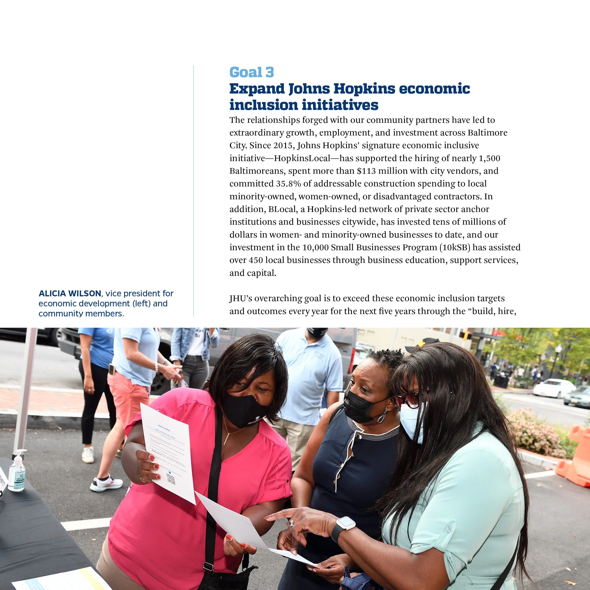 Three women wearing masks engaged in a discussion at an outdoor event, with printed materials in their hands and a table with hand sanitizer nearby, during daytime in an urban setting.