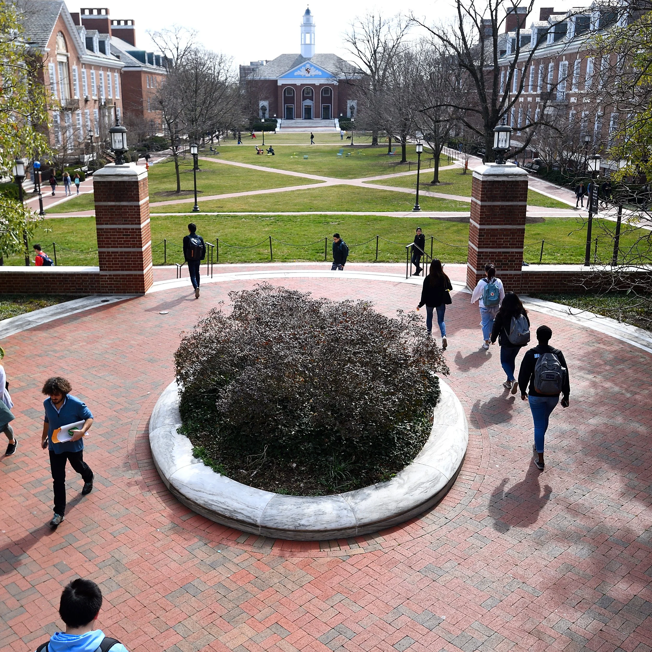 Students walking through a university campus with a brick plaza, a landscaped lawn, and a large building with columns and a clock tower in the background.