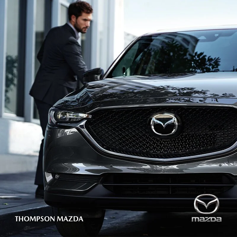 A man in a black suit is standing next to a black Mazda car outdoors, near a modern building with glass windows. The Mazda logo is prominently displayed on the front grille of the car.