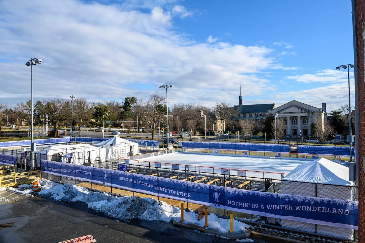 Outdoor ice skating rink with tents and snow, surrounded by a fence with winter-themed banners, in a city setting with buildings and trees in the background.