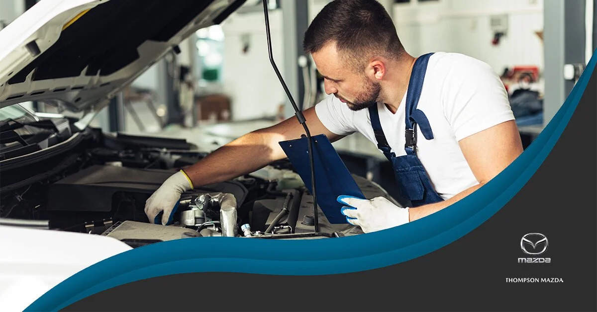 A male mechanic working on a car engine in a garage, wearing gloves and a white T-shirt with blue overalls, holding a clipboard.