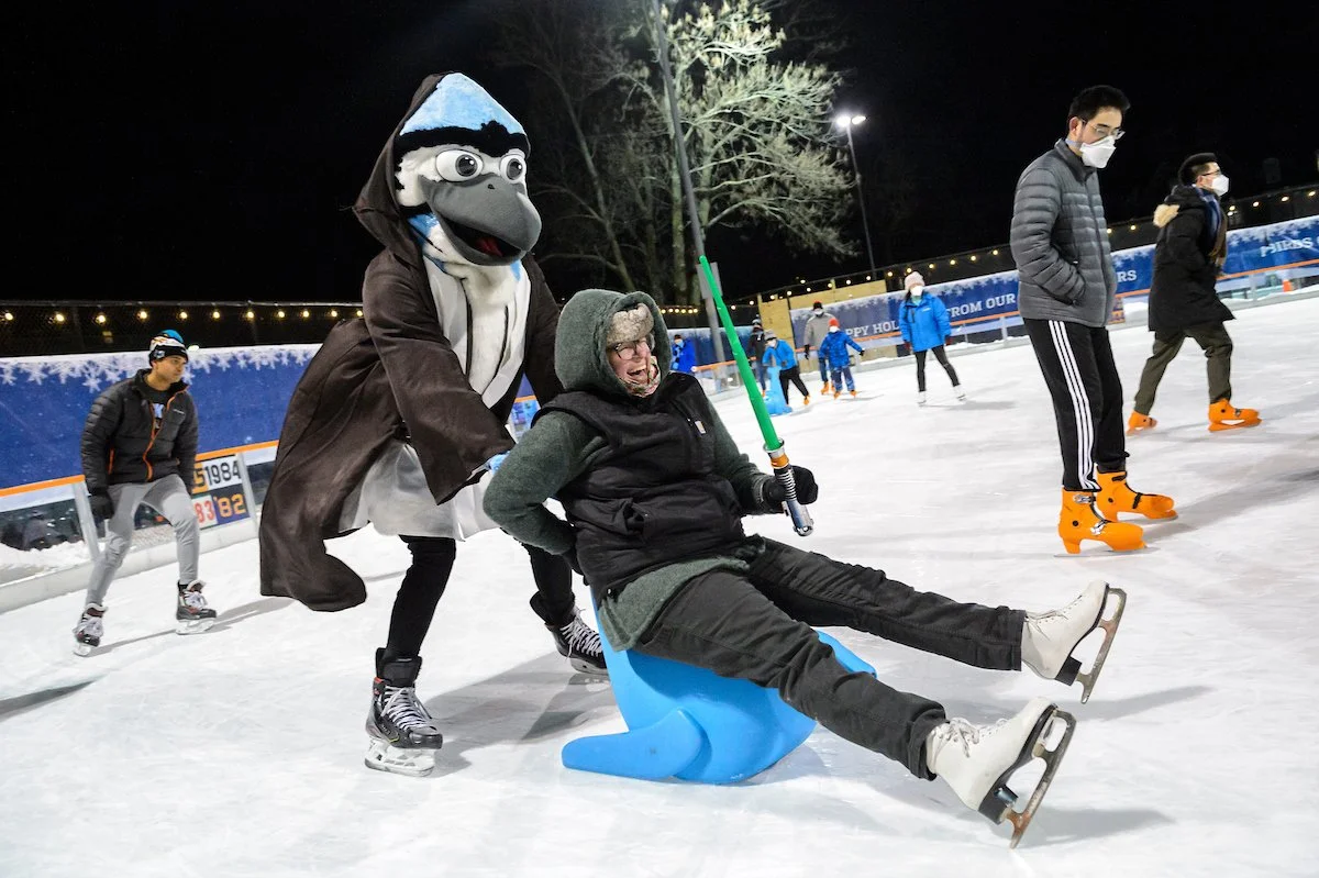 A person in a dolphin mascot costume pushing a seated person with a fish-shaped sled on an ice skating rink at night. Both are wearing winter clothing and ice skates.