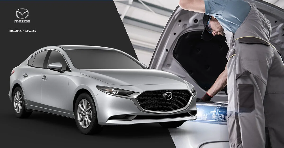 A silver Mazda sedan parked indoors next to a mechanic inspecting the car's engine with the hood open.