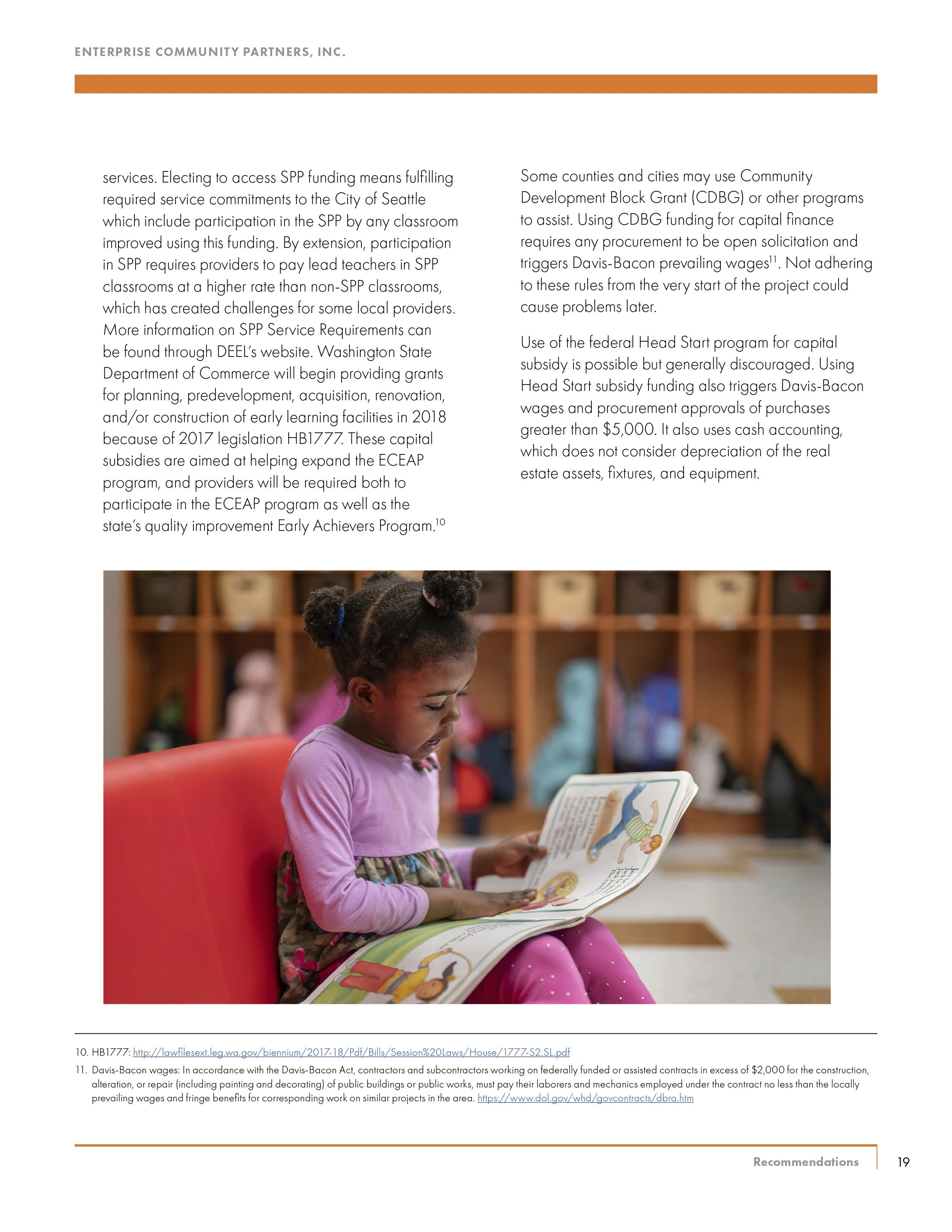 A young girl with braided hair and a pink long-sleeve shirt reading a children's book with colorful illustrations in a room with wooden cubbies filled with backpacks and other items.