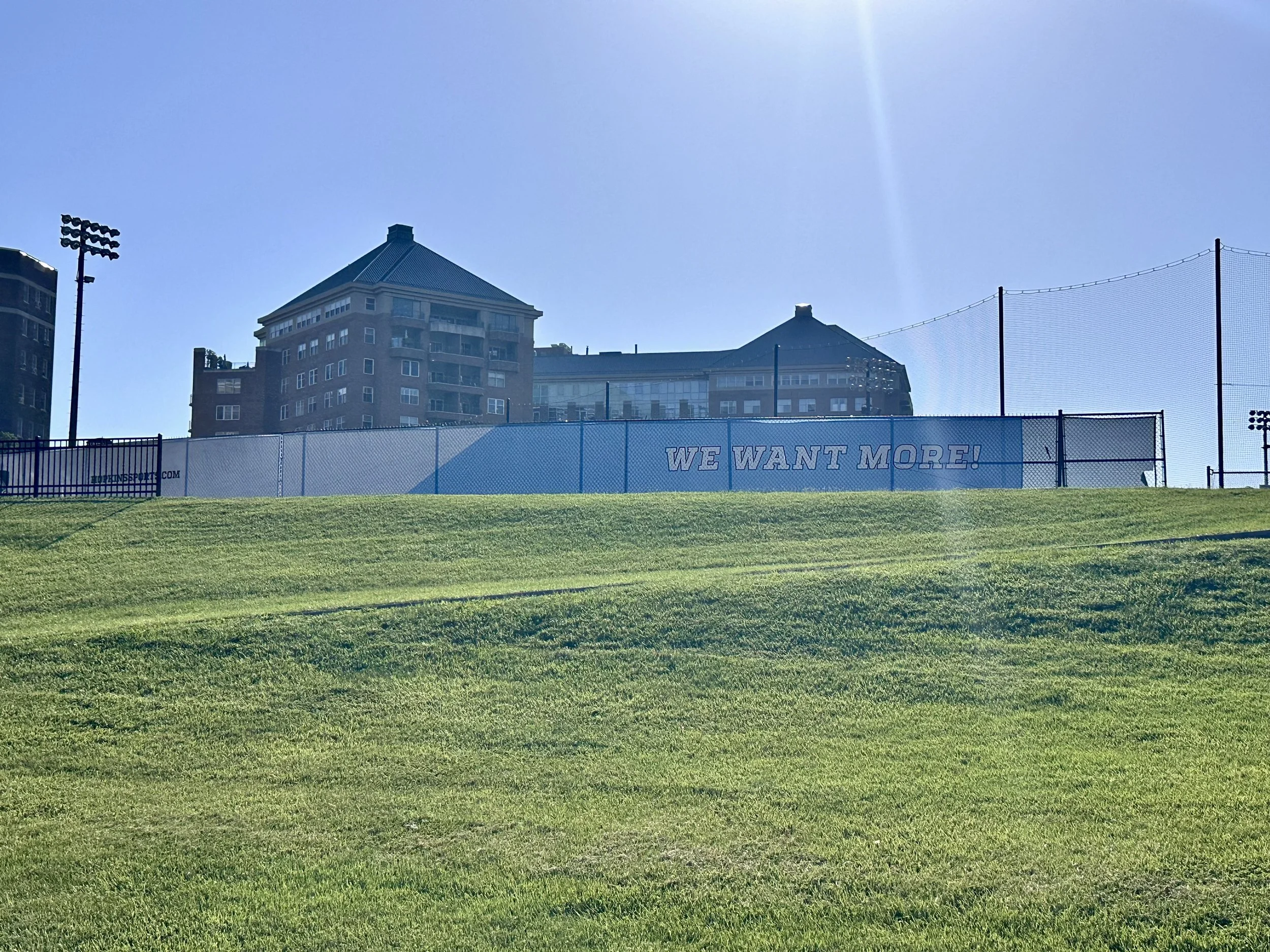 Green grassy hill with a sports field in the background, including a sign that reads 'WE WANT MORE!' with city buildings beyond and a clear blue sky.