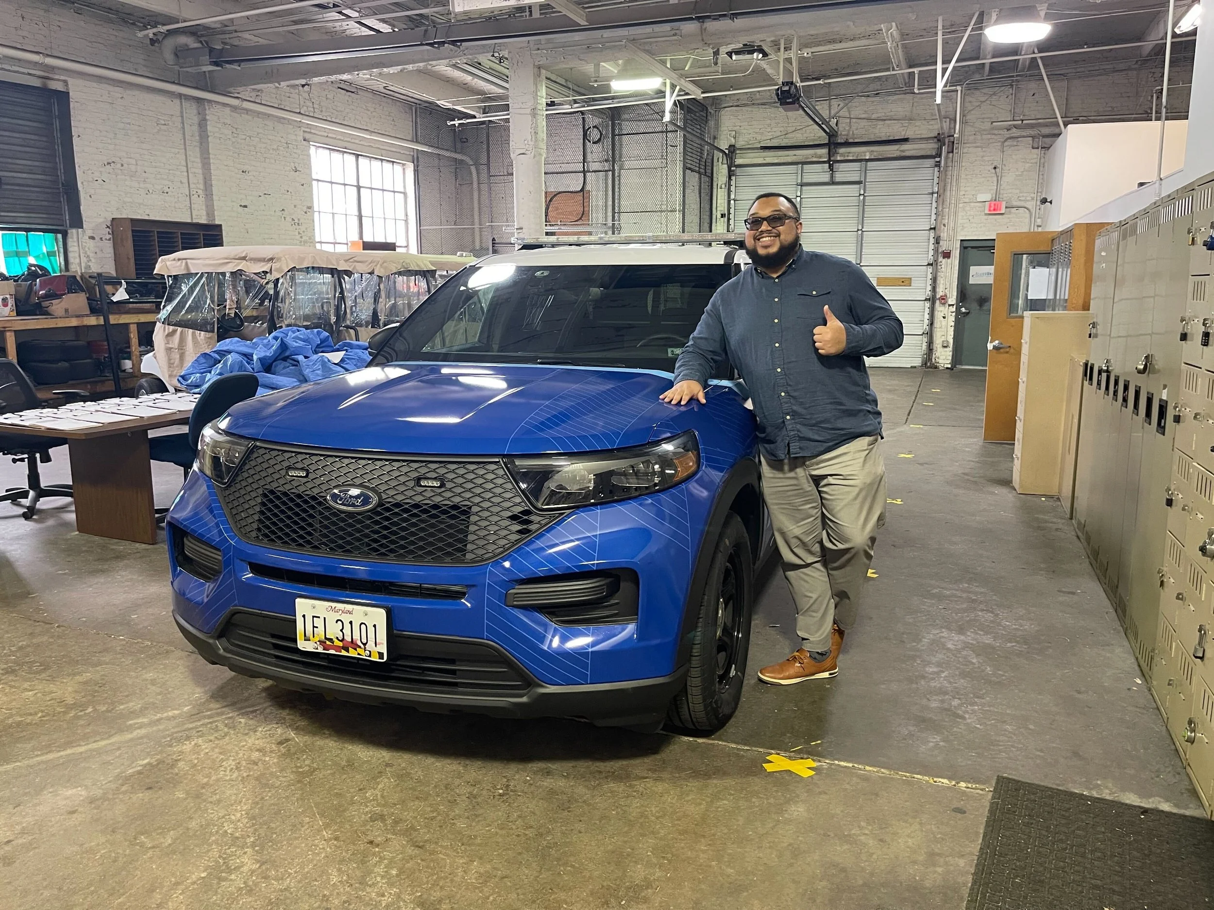 A man standing next to a blue Ford SUV inside an industrial garage, giving a thumbs up and smiling.