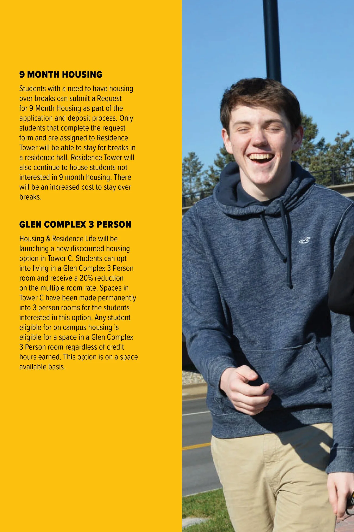 A young man laughing outdoors, wearing a blue hoodie and tan pants, standing on a sidewalk with trees and a blue sky in the background.