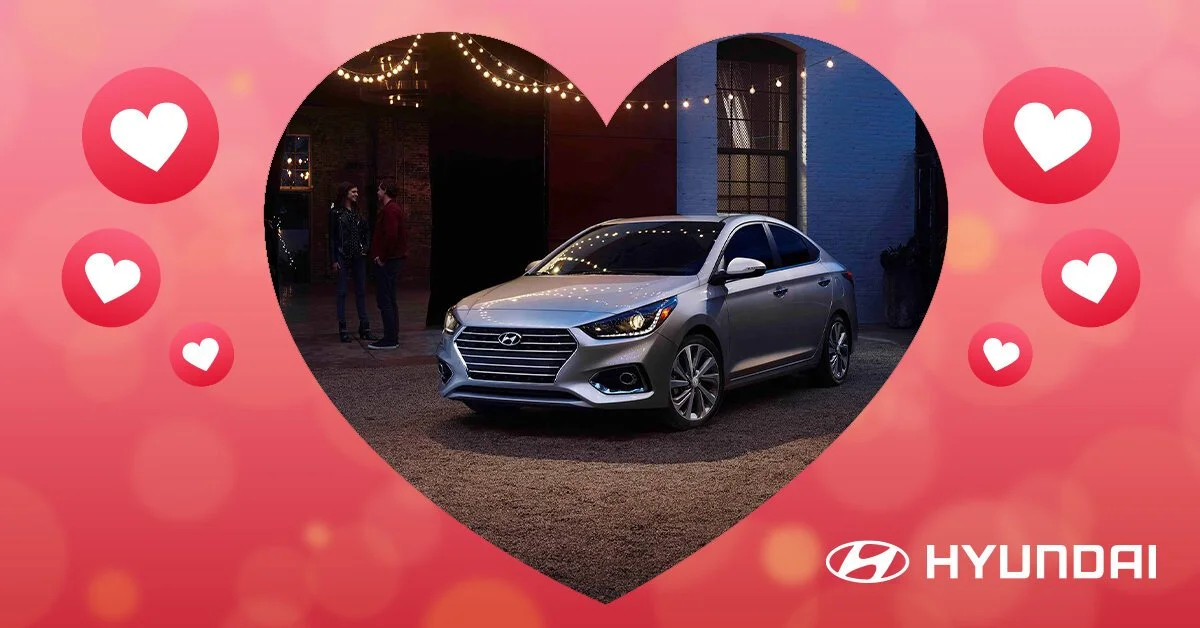 A silver Hyundai sedan parked inside a pink and red heart-shaped border with smaller hearts and the Hyundai logo, with a dark background and string lights overhead.