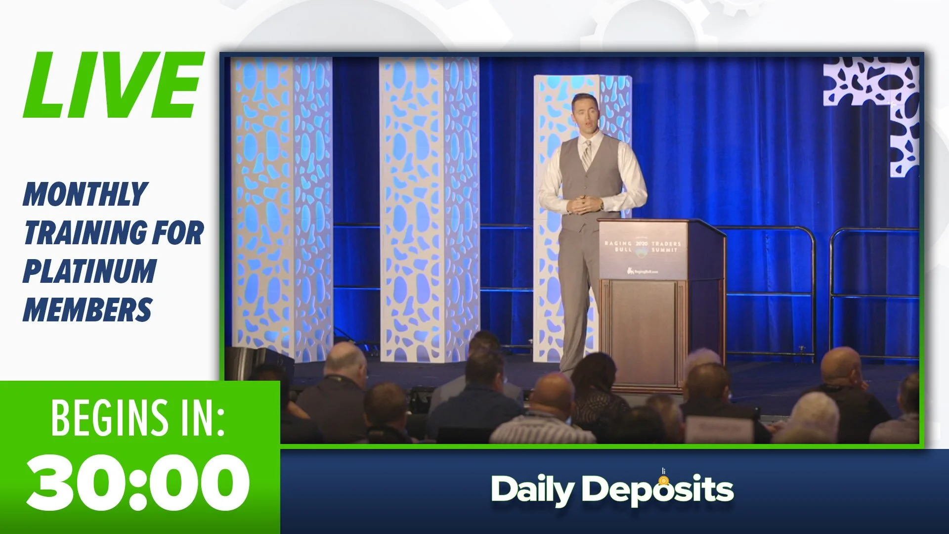 A man in a vest and tie standing at a podium on a stage with blue curtains and decorative panels, speaking to an audience at a conference or seminar.