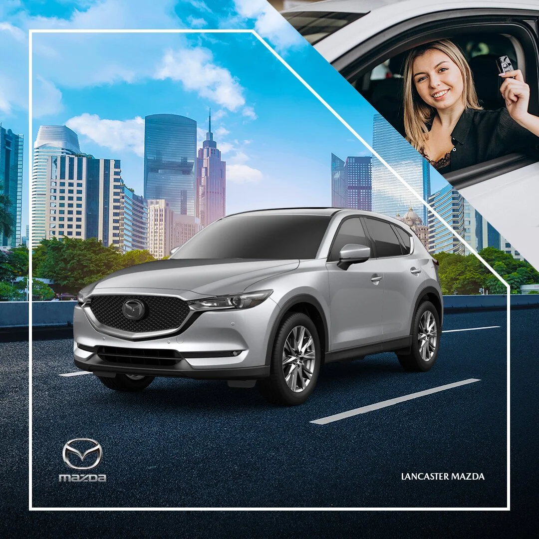 A silver Mazda SUV parked on a city street with tall buildings and blue sky in the background. A woman is smiling and holding a car key from the driver’s window.