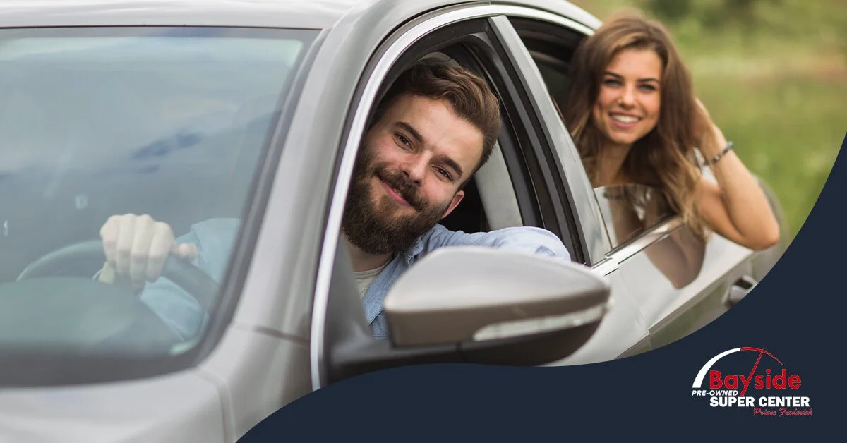 A smiling man with a beard leaning out of a passenger side window of a car, with a woman smiling in the background, outdoors near a body of water.
