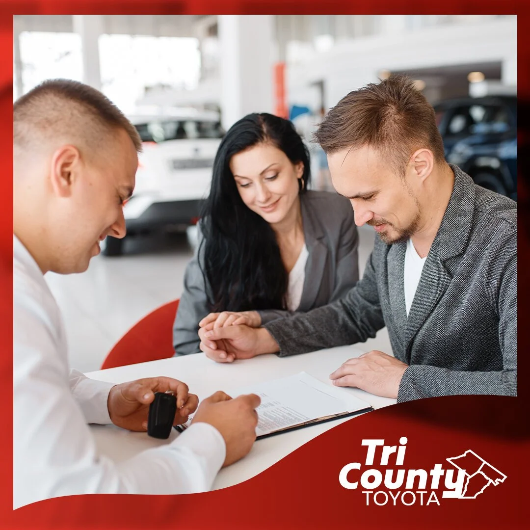 A couple is sitting with a car salesperson at a dealership, discussing paperwork, with cars in the background and Tri County Toyota logo in the corner.