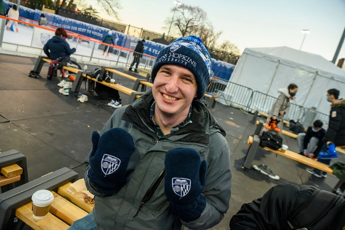 Smiling man in winter clothing holding up navy gloves with a logo at an outdoor ice skating rink.