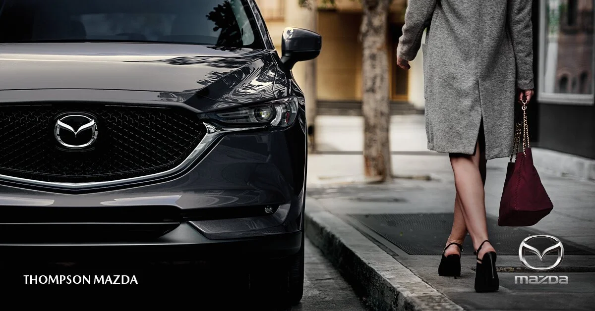 A woman walking on a city sidewalk near a parked black Mazda car, wearing a gray coat and high heels, carrying a maroon bag.