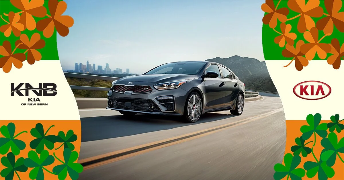 A gray Kia KNB sedan driving on a highway with mountains in the background, framed by graphic St. Patrick's Day-themed clovers and banners featuring Kia logos.