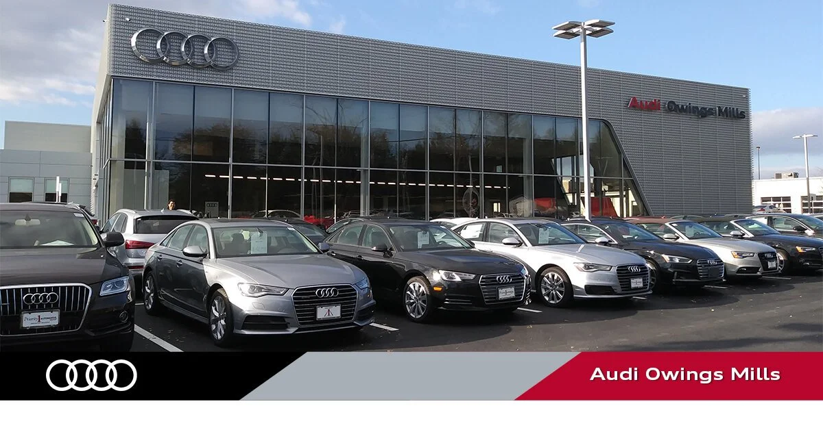 Exterior view of Audi dealership with several Audi cars parked in front, large glass windows, and an Audi logo on the building. Sign reads 'Audi Owings Mills'.