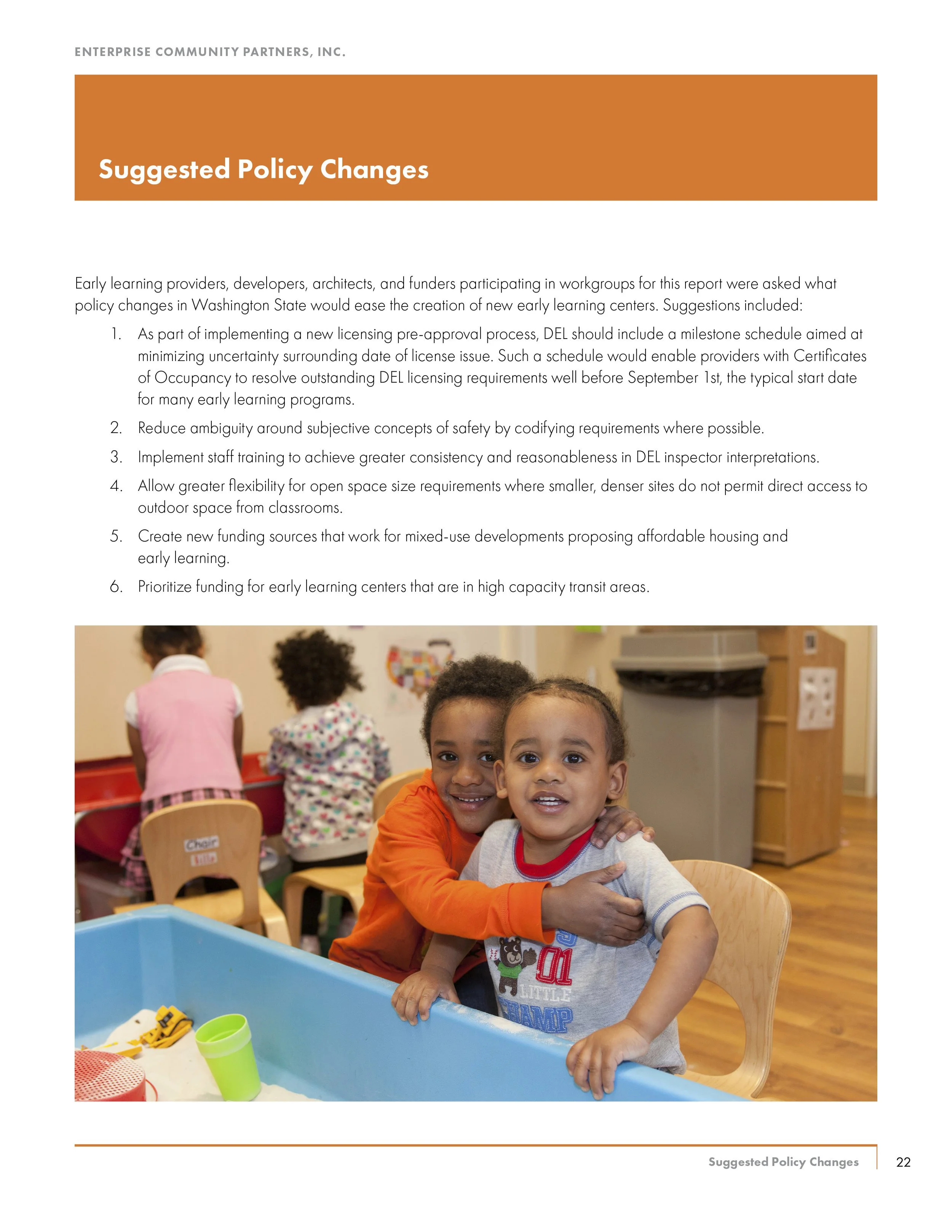 Two young children, one with a big smile, hugging each other while sitting at a table in a classroom. Other children are visible in the background, engaged in activities.