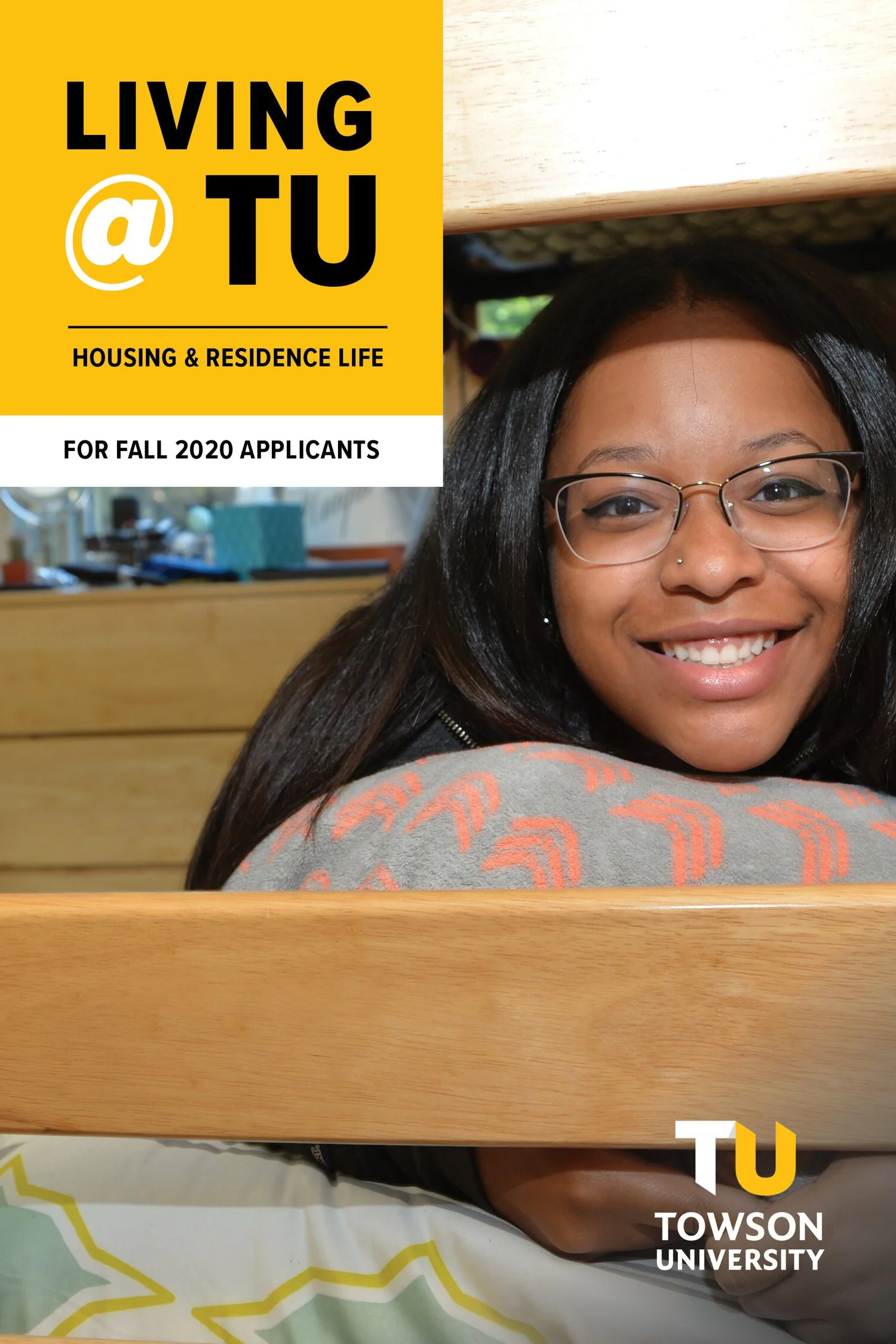 A young woman with glasses and a nose piercing smiling at the camera, sitting at a wooden table in a university dining area, with promotional text overlay for Towson University housing applications for fall 2020.