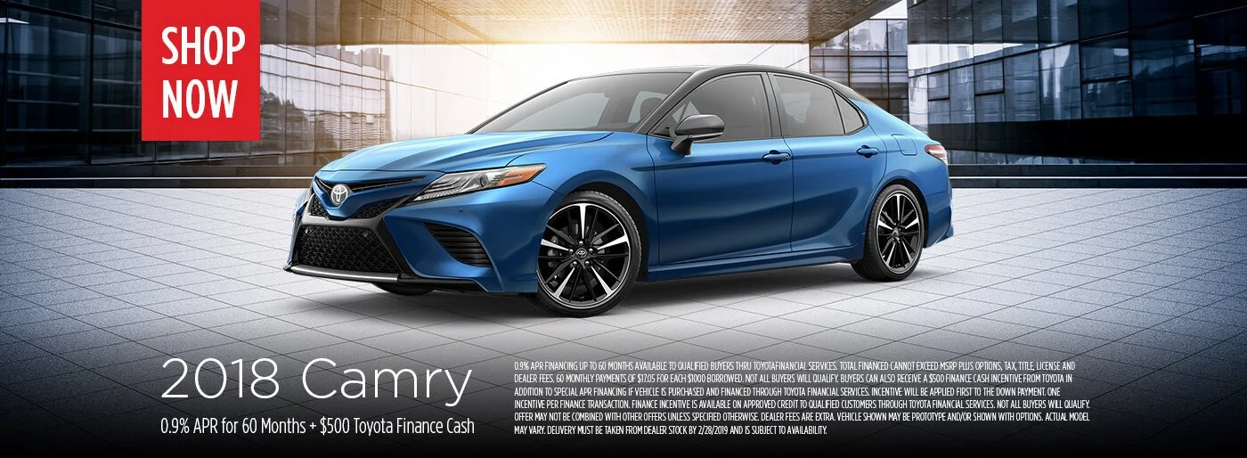 A blue 2018 Toyota Camry is displayed in a modern showroom with glass walls and a tiled floor. There is a red rectangle with the text 'Shop Now' in the upper left corner.