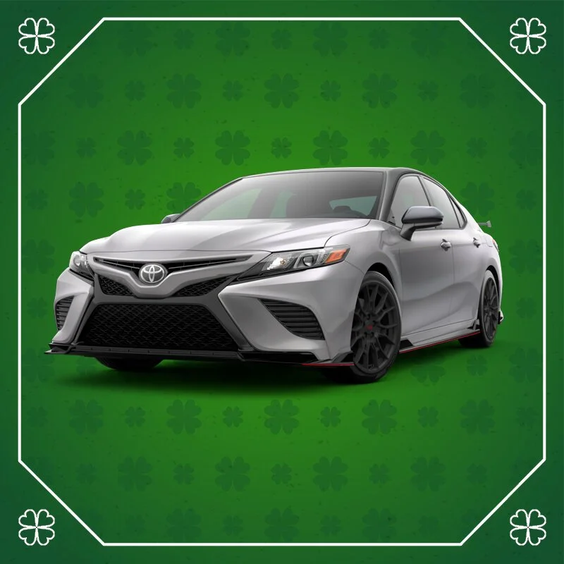 A silver Toyota Camry sedan with black wheels, front grille, and side mirrors, positioned on a green background with shamrock patterns and white border.