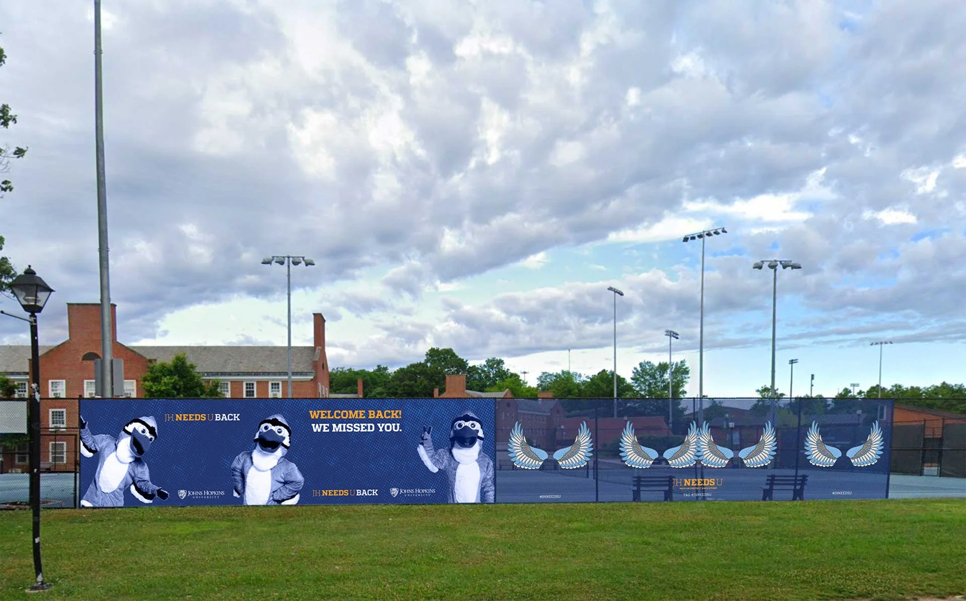 A blue banner with images of a mascot, a winged emblem, and text, on a tennis court with a grassy area in the foreground, trees, and residential buildings with brick facades in the background under a cloudy sky.