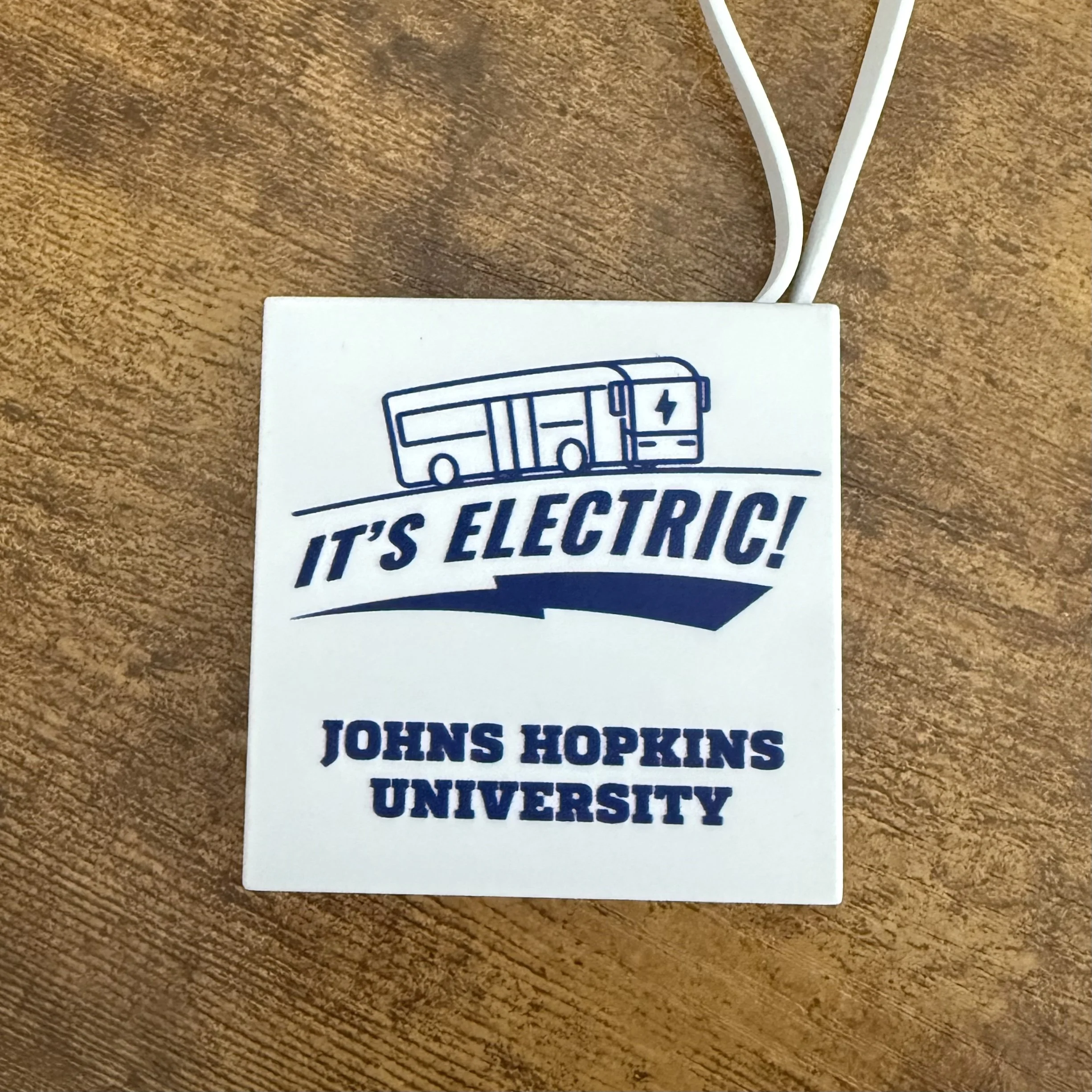 Square white device with a blue graphic of an electric bus and the text 'IT'S ELECTRIC! JOHN HOPKINS UNIVERSITY' printed on it, placed on a wooden surface. Two white cords are attached to the device.