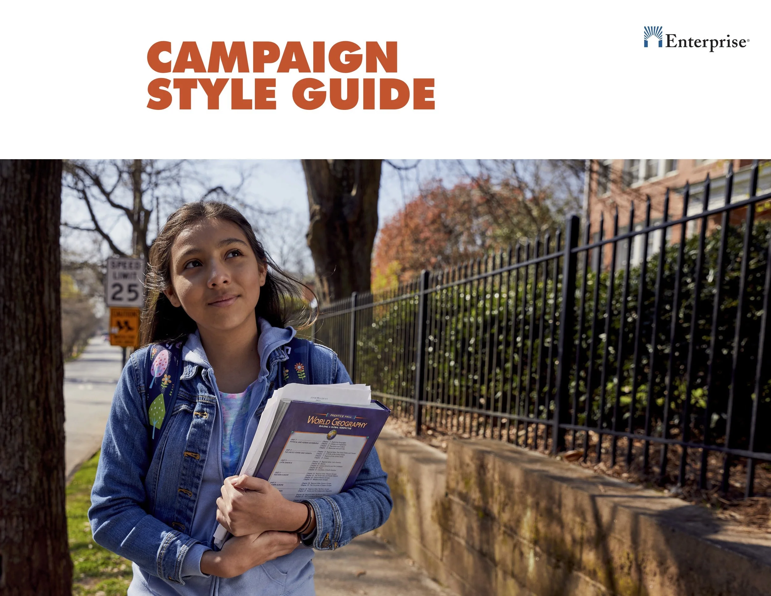 A young girl holding textbooks walk along a sidewalk with trees and a black metal fence in the background on a sunny day.