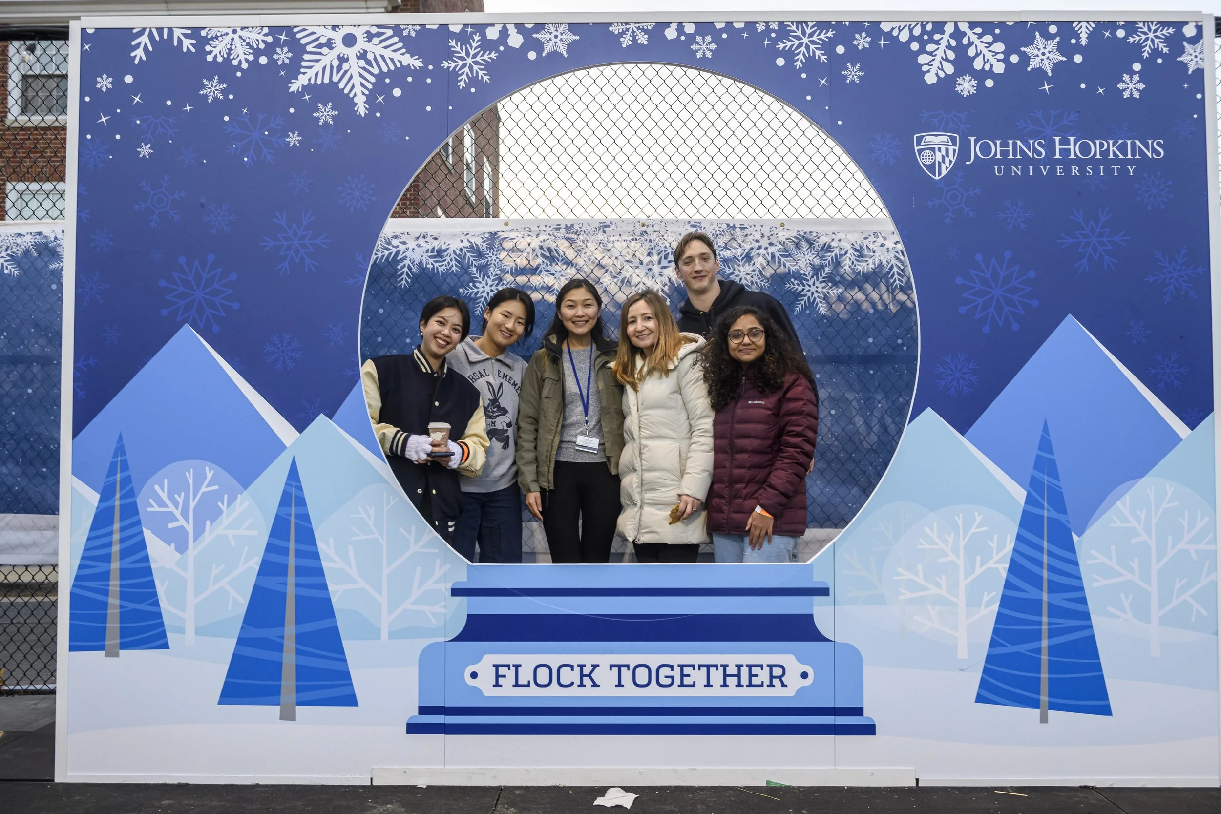 Group of six diverse people standing behind a winter-themed photo booth with a snowy mountain and pine trees design, displaying the Johns Hopkins University logo and the words "Flock Together".