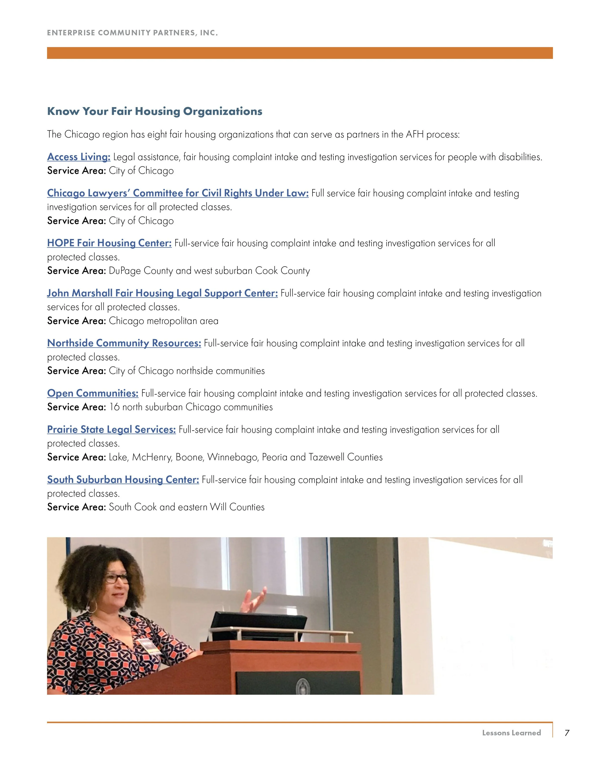 A woman with curly hair and glasses giving a presentation at a podium, with a projection screen in the background.