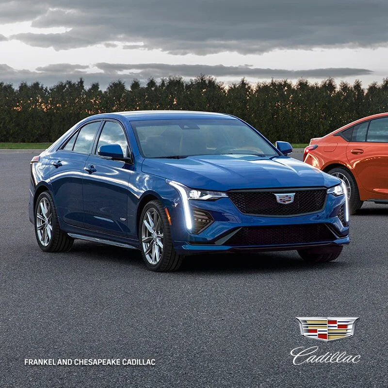 A blue Cadillac sedan parked on a paved surface with another orange car in the background and dark cloudy sky above.
