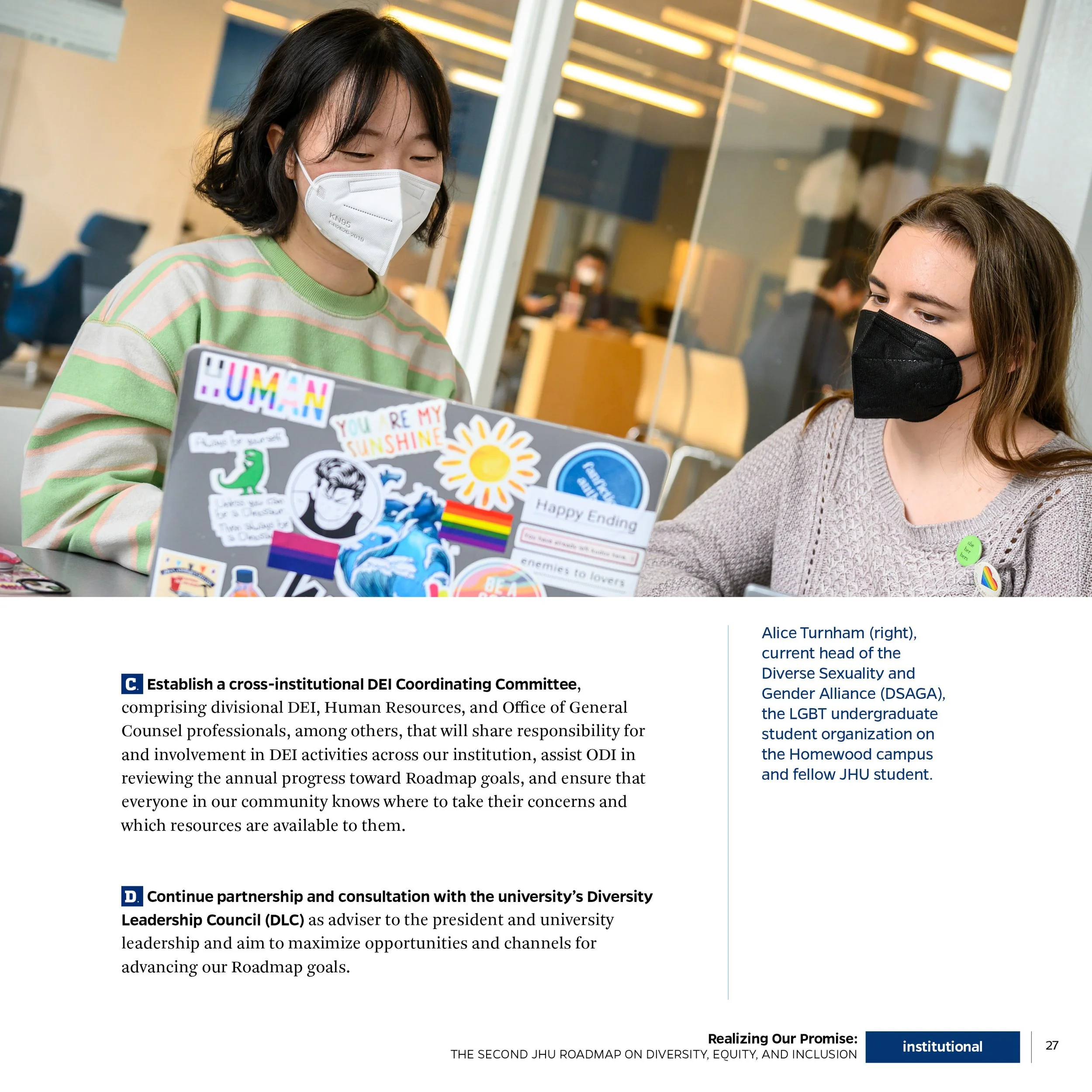 Two women wearing masks and sitting at a table with a laptop decorated with stickers, inside a room with glass walls and people in the background.