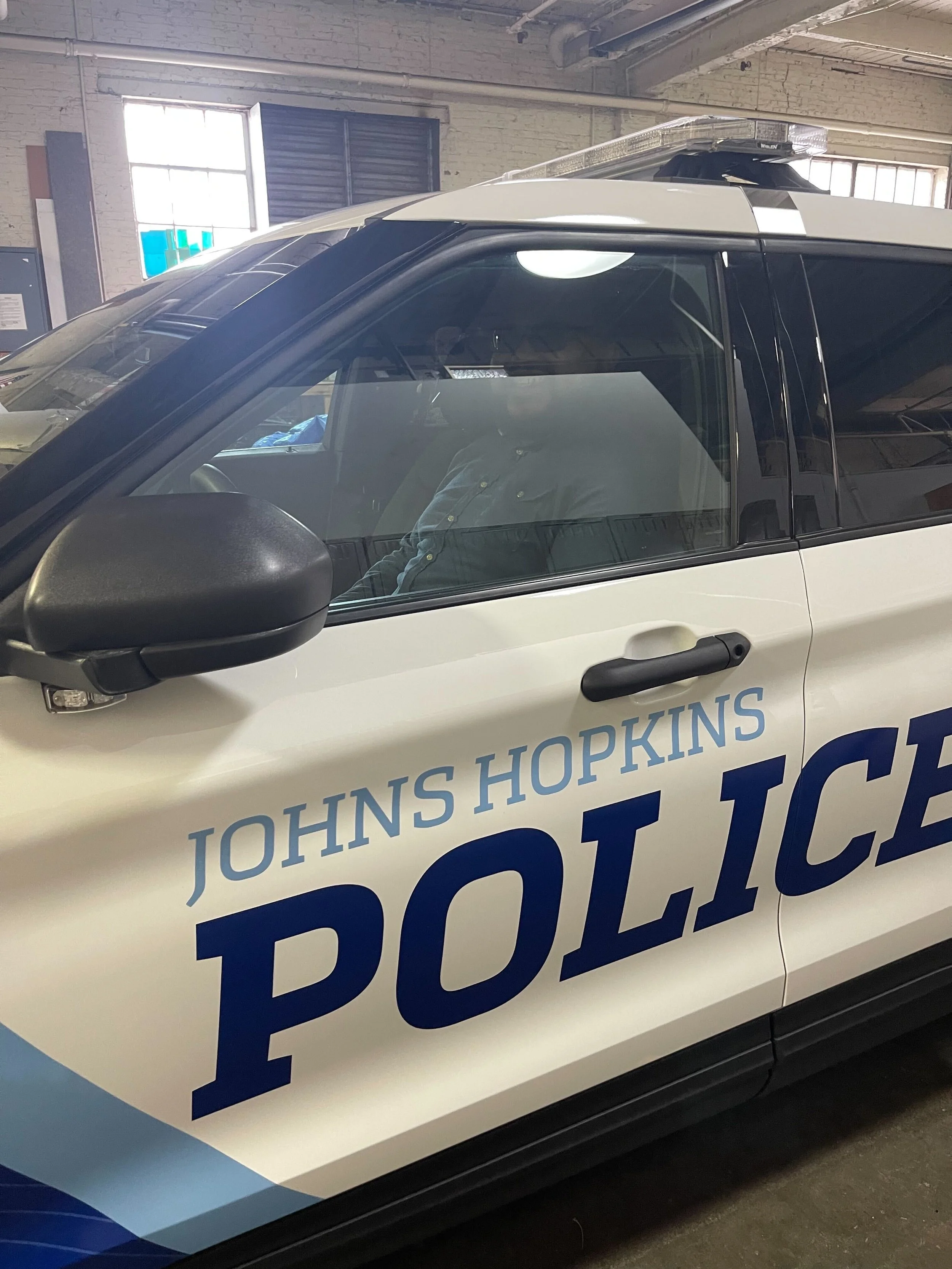 A police car from Johns Hopkins, inside a garage or indoor parking area, with a person visible through the window.