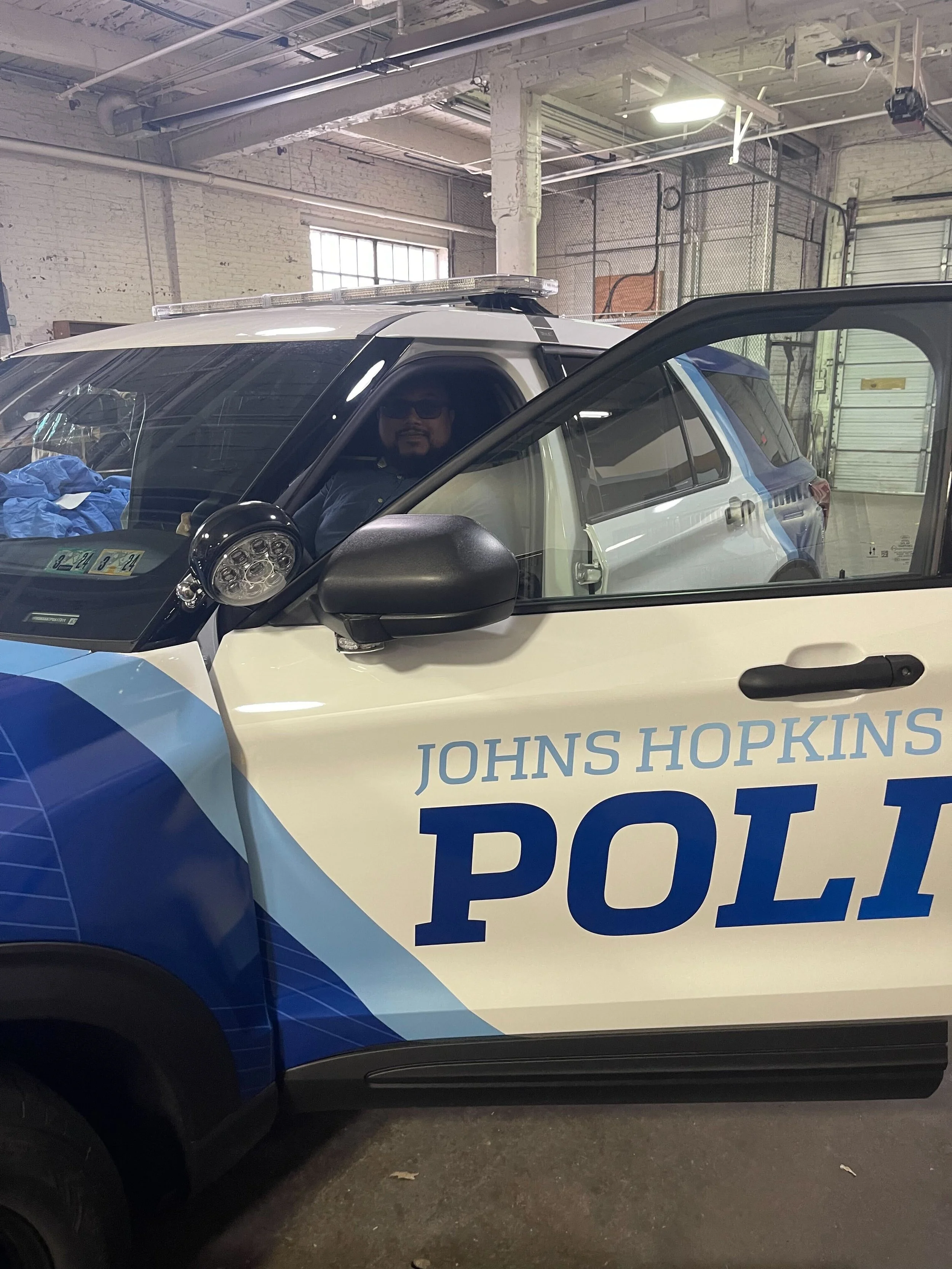 A police vehicle with 'Johns Hopkins POLI' on the side, parked in an indoor garage. A police officer is sitting inside the vehicle, visible through the open driver’s side window.
