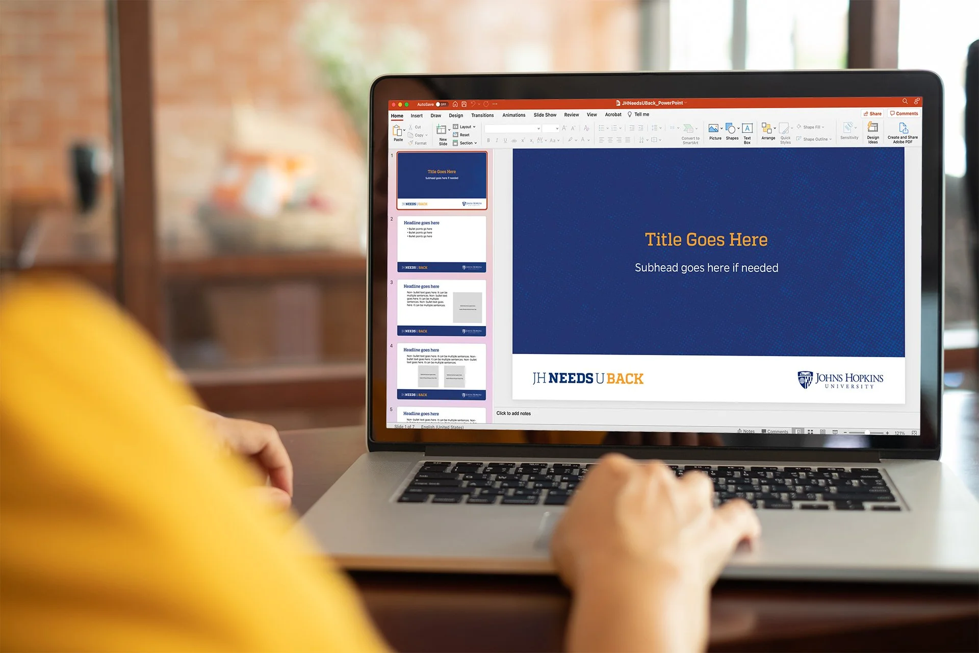 A person working on a laptop displaying a PowerPoint presentation with the title "JH NEEDS U BACK" and the Johns Hopkins University logo, in a room with a brick wall and large window in the background.