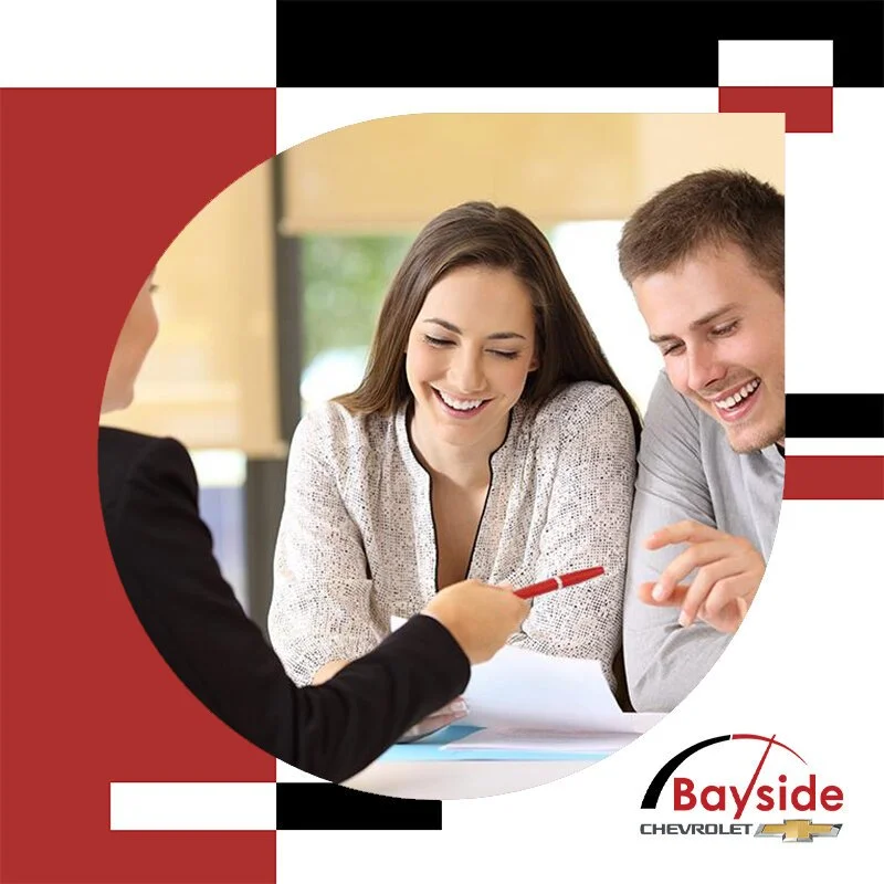 A woman and a man smiling and looking at a woman who is showing them something with a red pen, in a meeting room with bright natural light, with a Bayside Chevrolet logo at the bottom.