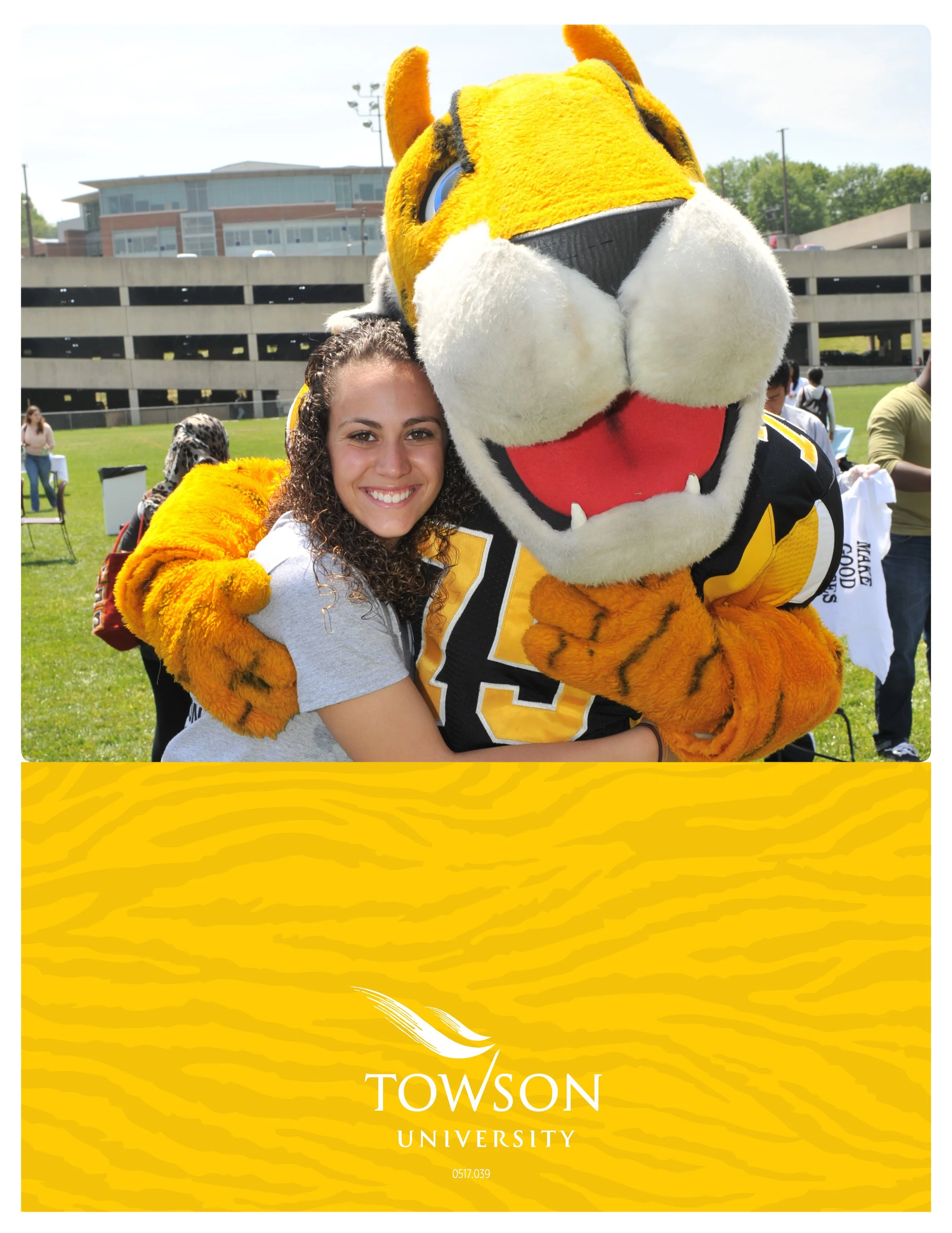 A young woman smiling and hugging a mascot dressed as a tiger in a sports jersey with the number 13. The background shows a grassy field, a modern building, and people gathered outdoors on a sunny day. The bottom of the image has a yellow section wit