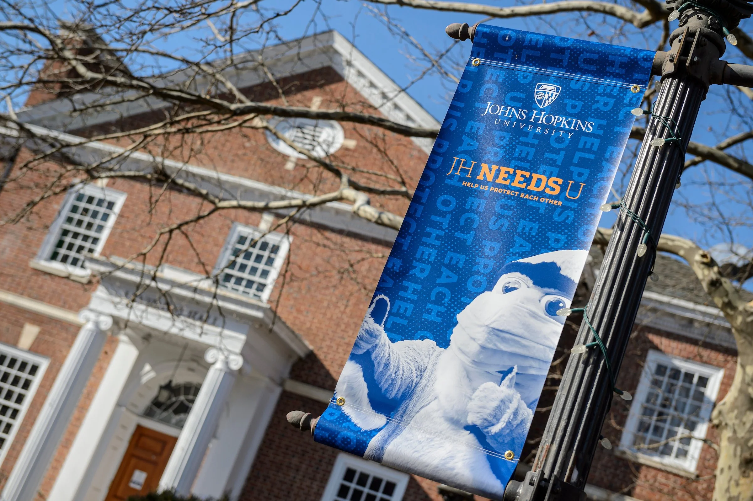 Blue Johns Hopkins University banner with a snowman wearing a face mask giving a thumbs up, hanging on a black pole in front of a brick building with white columns and windows, and a tree with bare branches against a blue sky.