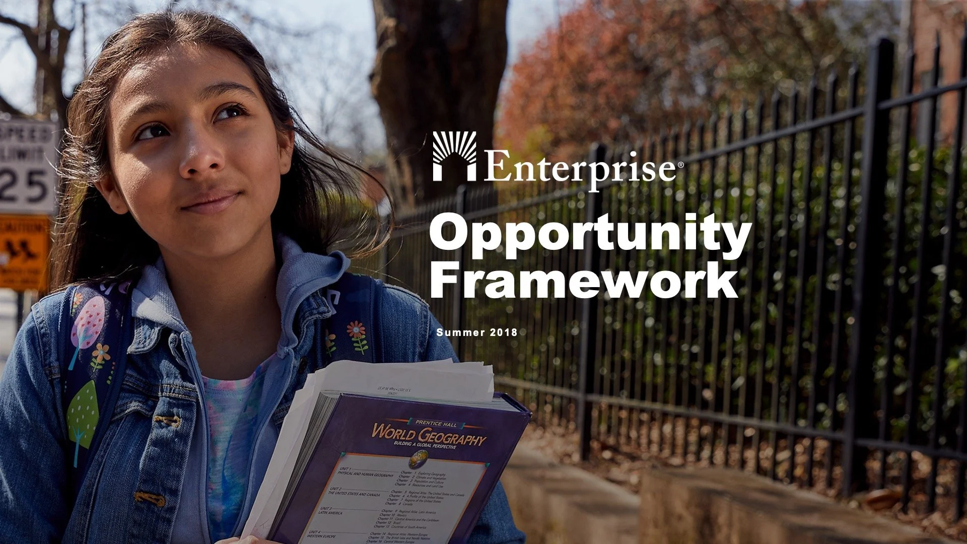 A young girl with long brown hair, wearing a denim jacket and carrying a backpack, holding a world geography textbook, standing outside near a black metal fence with trees in the background, on a sunny day.
