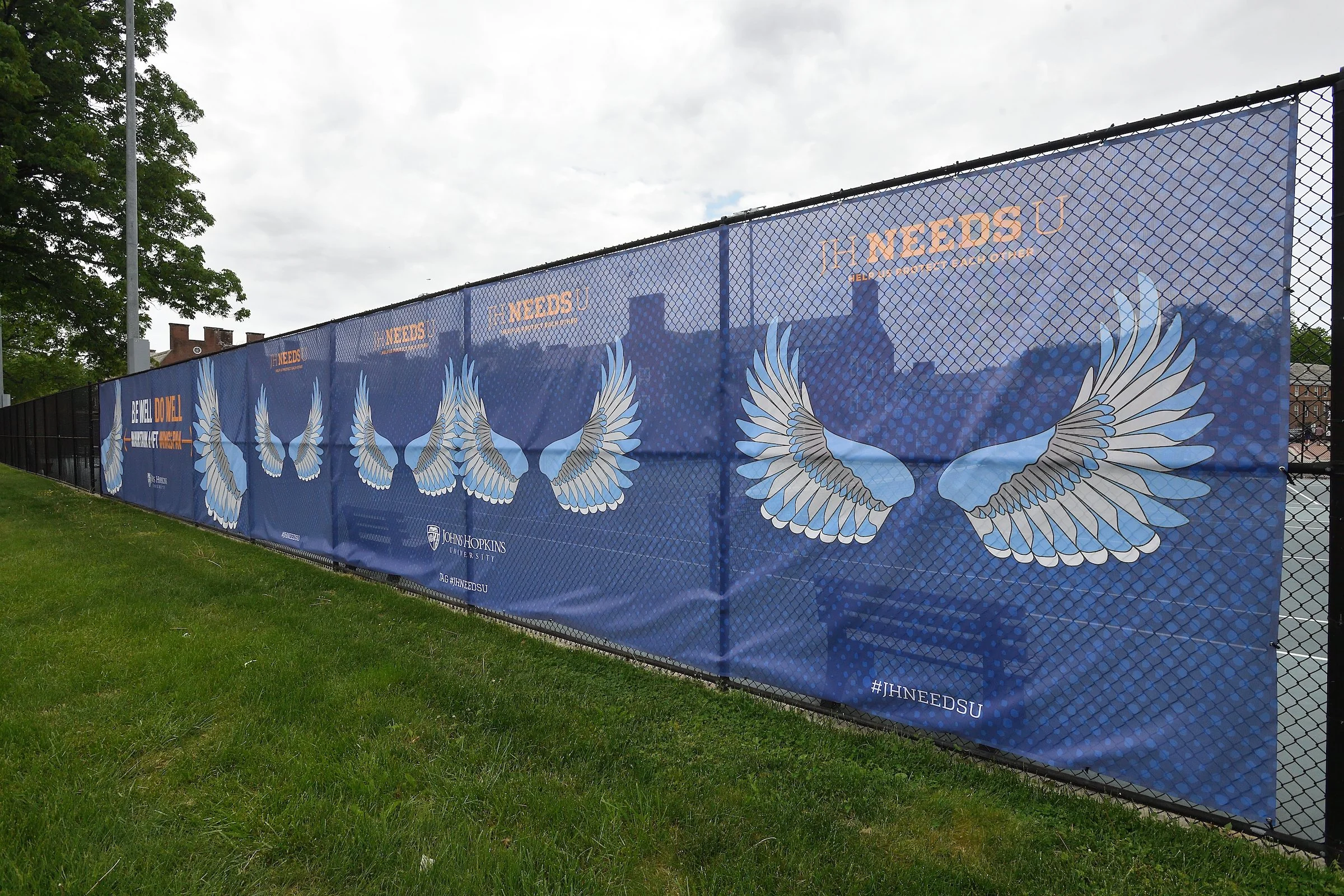 Banner on a chain-link fence featuring angel wings illustrated with shades of blue and white, and text promoting Johns Hopkins University. The background shows a cloudy sky and some trees.