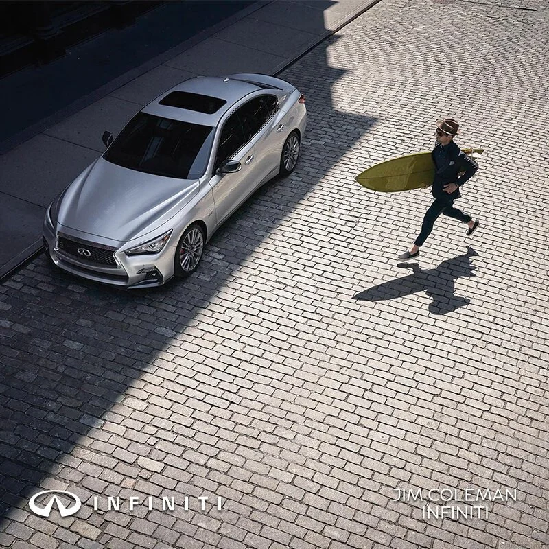 A silver Infiniti sedan parked on a cobblestone street, with a woman running past it carrying a surfboard, casting a large shadow. Logos and text in the lower corners read 'INFINITI' and 'JIM COLEMAN INFINITI'.