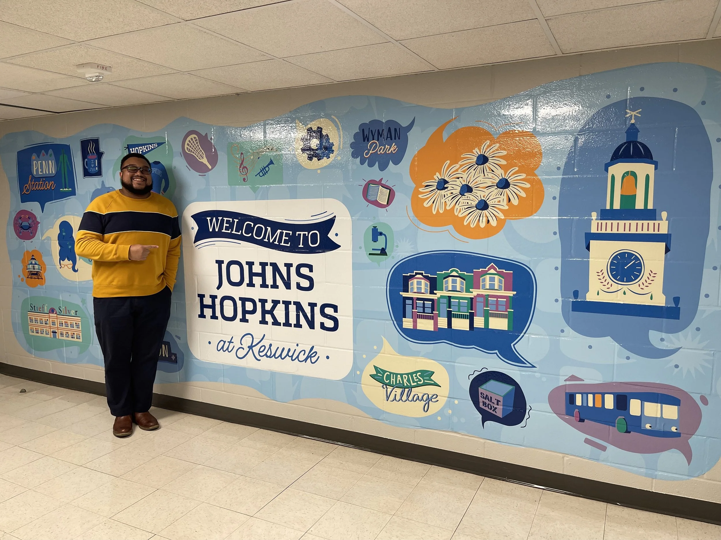 A man standing in front of a colorful mural that says 'Welcome to Johns Hopkins at Keswick' with illustrations of city landmarks, flowers, a clock tower, and various icons representing activities and places in the area.