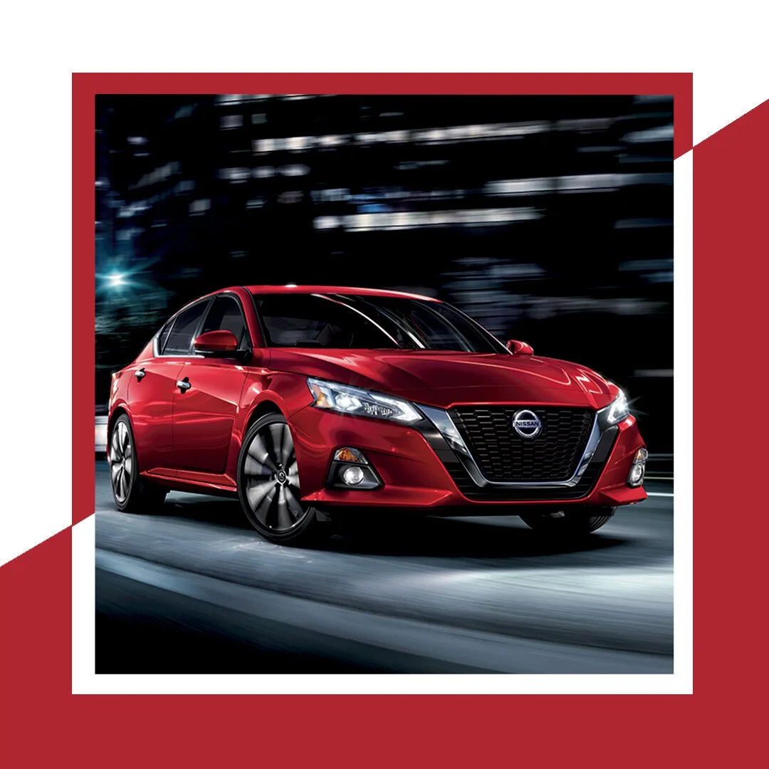 Red Nissan sedan driving on a city street at night with blurred lights in the background.