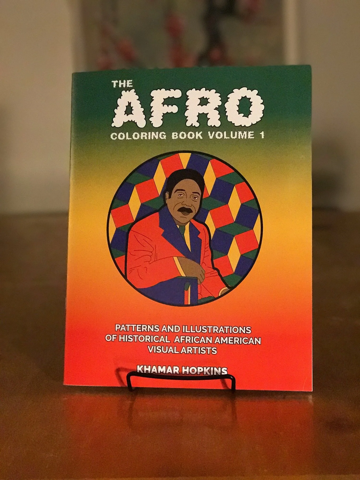 Cover of a coloring book titled "The Afro Coloring Book Volume 1" featuring an illustration of an African American man in a red and blue outfit with a colorful geometric background, and text about patterns and illustrations of historical African Amer